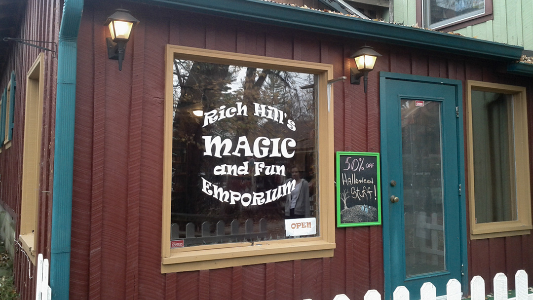 Exterior of a red building with a turquoise door with a sign in the window reading "Rich Hill's Magic and Fun Emporium