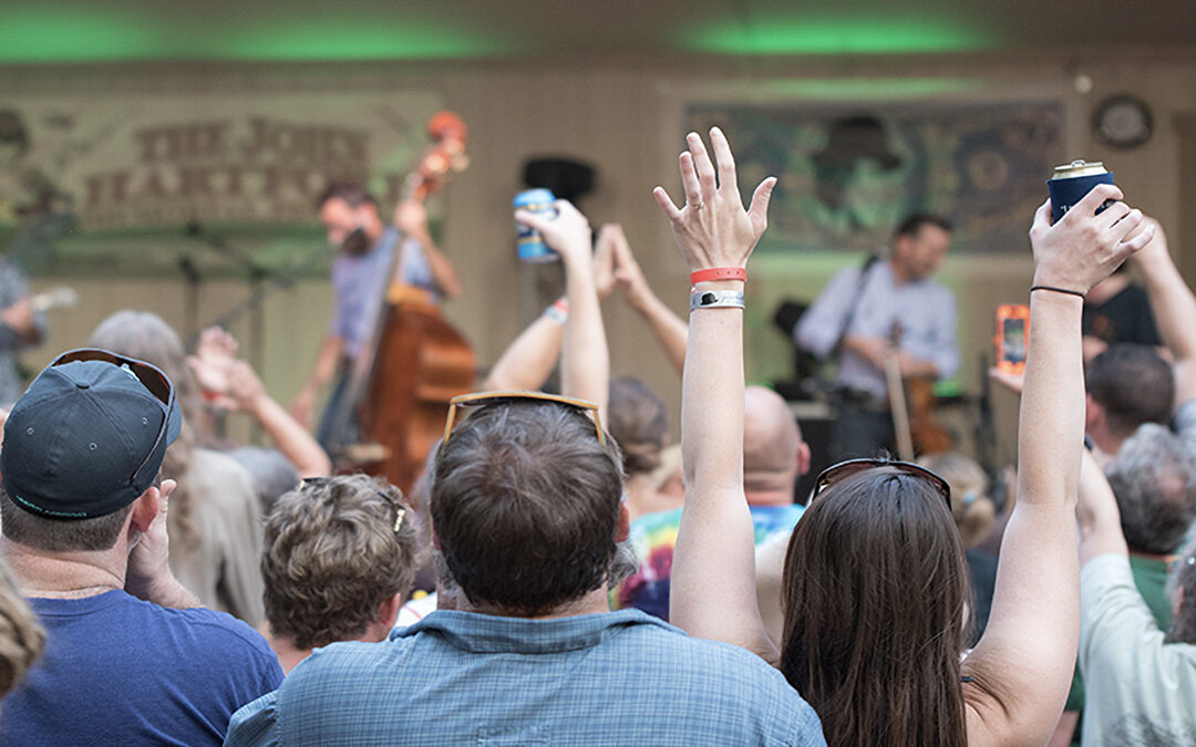 hands are raised in the foreground as a crowd watches bluegrass performers on stage