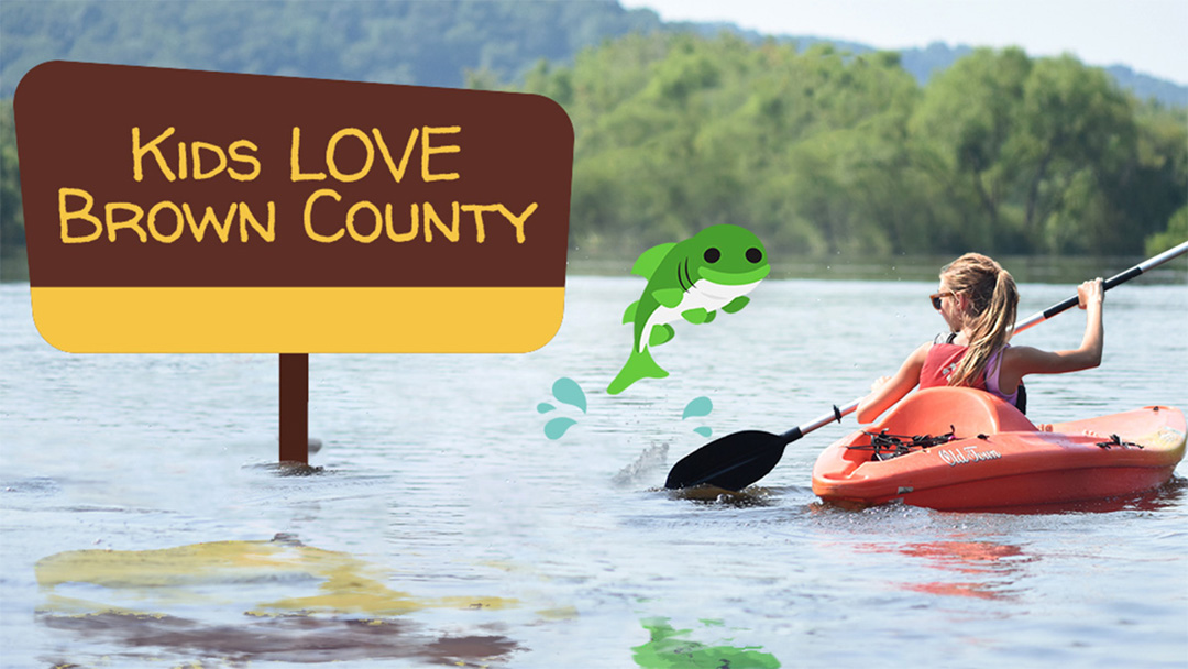 Young girl kayaking while a cartoon fish jumps out of the water near a cartoon sign which reads "Kids LOVE Brown County"