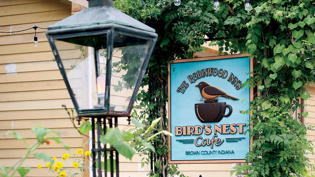 Old fashioned street light in the foreground with a blue Bird's Nest Cafe sign, trellis and greenery behind it