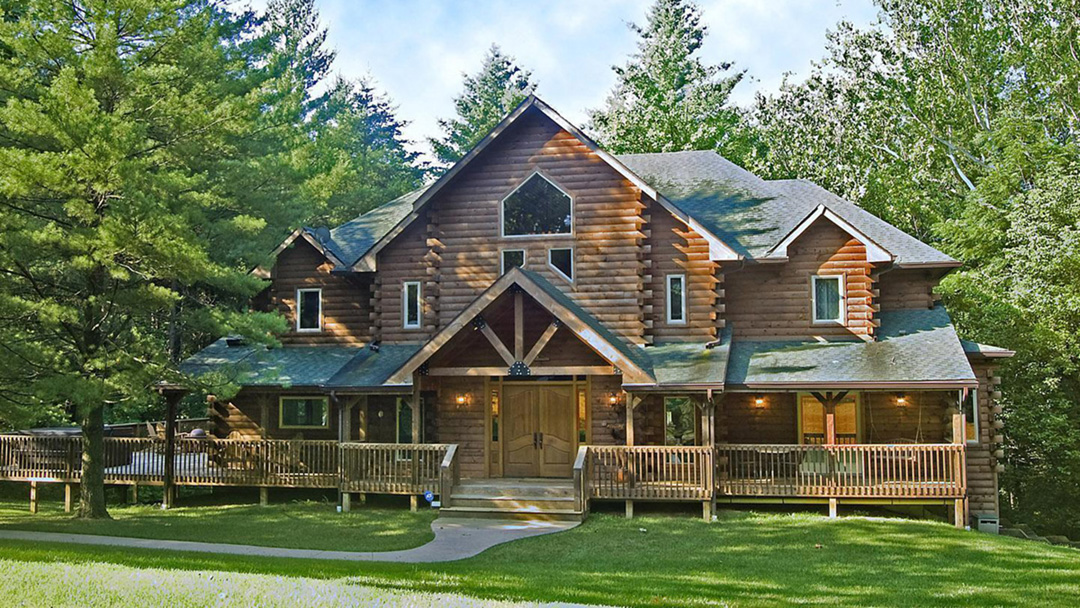 Exterior of a large log cabin with an expansive porch and lush green lawn with trees behind the cabin