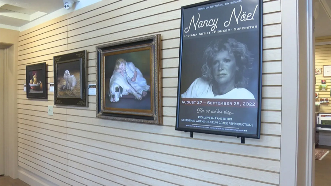 A row of three Nancy Noel paintings are hung on the wall of an art gallery with a poster advertising the exhibit in the foreground.