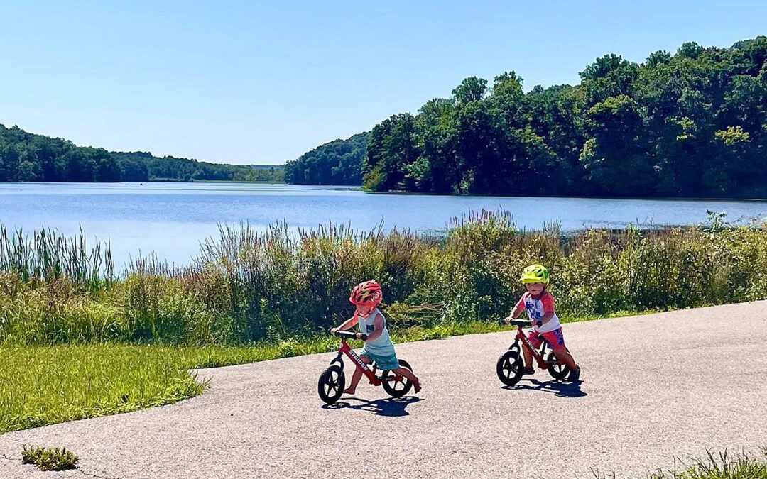 two young children riding bikes on a path with a tree lined lake in the background